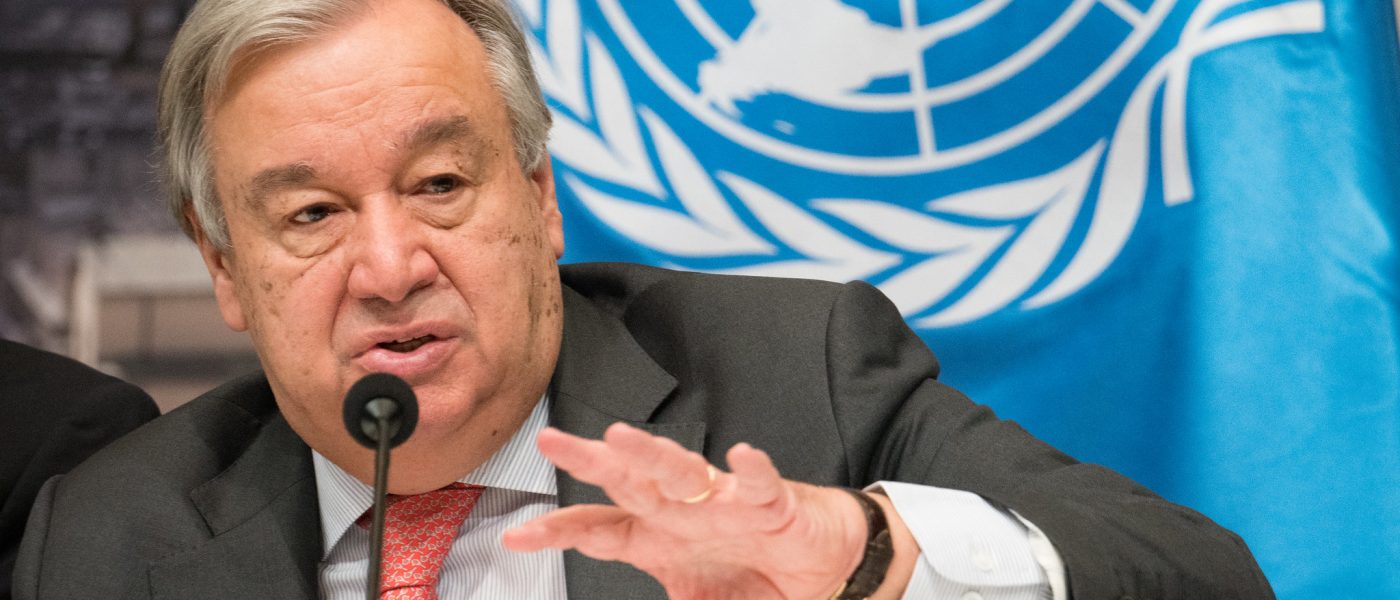 Mr. António Guterres, Secretary-General of the United Nations, speaking into a microphone in front of the UN flag.