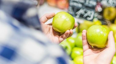A man in a grocery store considering two green apples he is holding in his hands