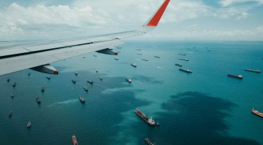 An airplane wing over a sea dotted with many cargo ships in the daytime